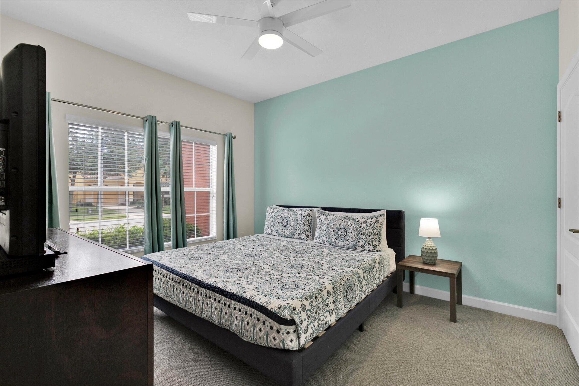 A bedroom with a bed, a nightstand, a lamp, a ceiling fan, and a window with curtains.