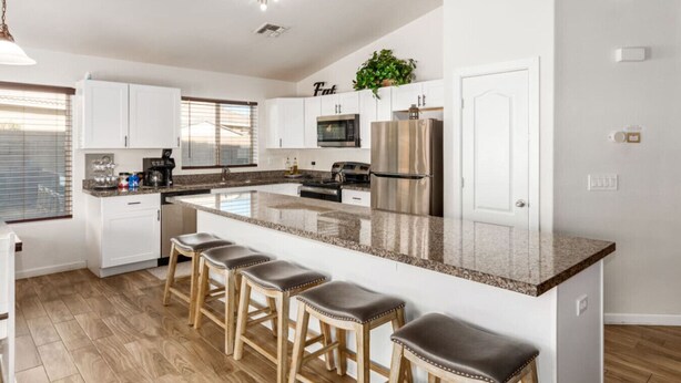 A modern kitchen with white cabinets, a stainless steel refrigerator, a microwave, and a large island with bar stools. - Beautifully Decorated Homeavondaleprivatepool (Avondale)