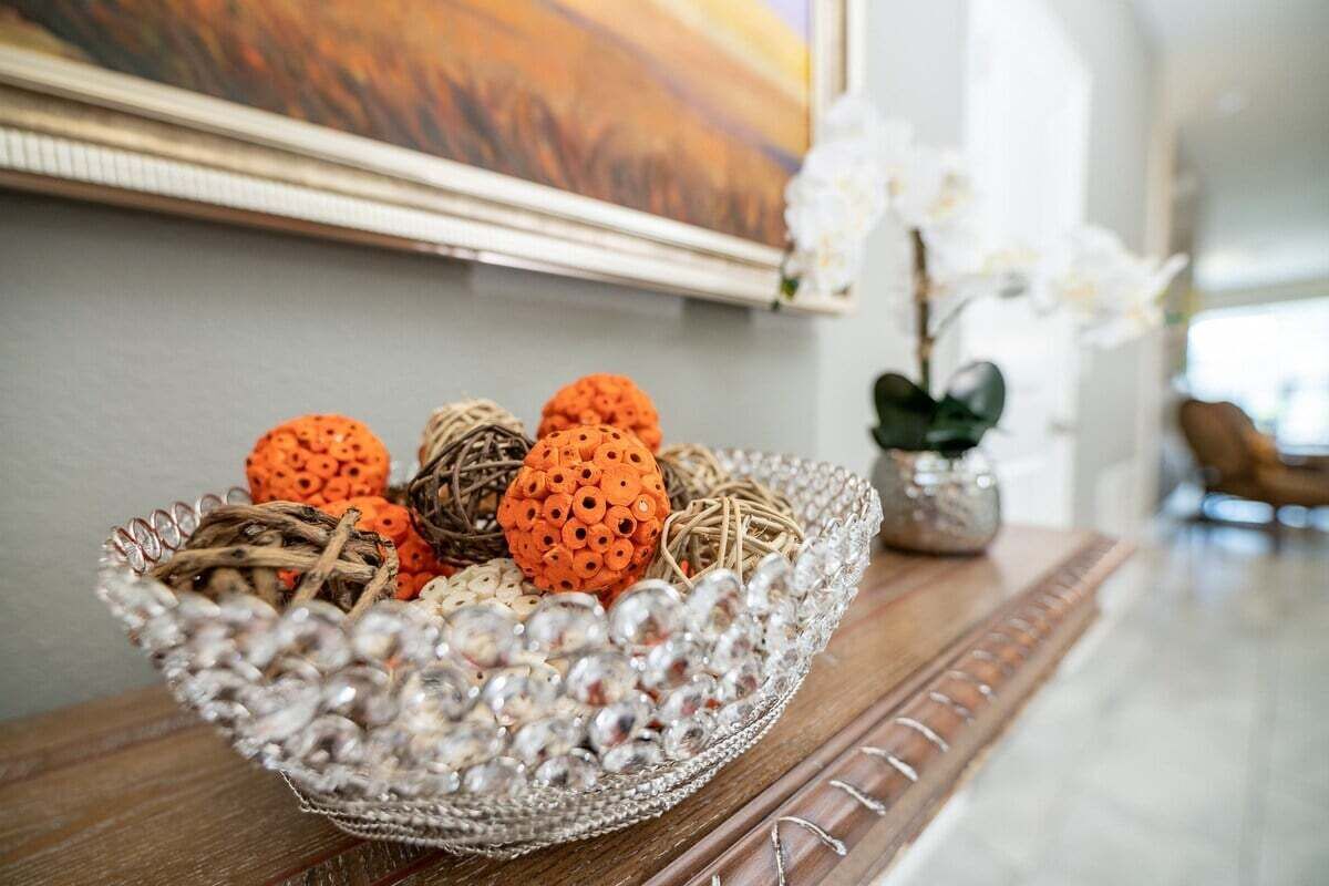 Eine dekorative Glasbowl mit orangefarbenen und weißen, kugelförmigen Objekten auf einem Holztisch, im Hintergrund ein Gemälde und weiße Blumen.