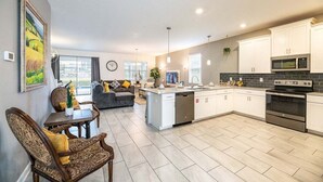 A modern kitchen with white cabinets, a built-in microwave, and a stainless steel oven. There is a dining area with a wooden table and chairs, and a sofa in the background. - Dazzing 9BR Pool Spa Free Waterpark (Davenport)