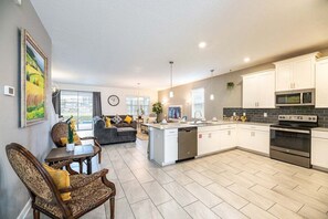 A modern kitchen with white cabinets, a built-in microwave, and a stainless steel oven. There is a dining area with a wooden table and chairs, and a sofa in the background. - Dazzing 9BR Pool Spa Free Waterpark (Davenport)