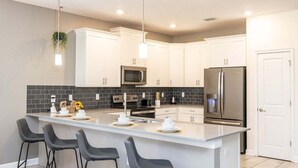 A modern kitchen with white cabinets, a central island with bar stools, stainless steel appliances, and a black tiled backsplash. - Dazzing 9BR Pool Spa Free Waterpark (Davenport)