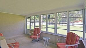 A covered porch with two striped chairs and a small table, large windows offering a view of a yard and trees. - Beechwood Villa (Sarasota)