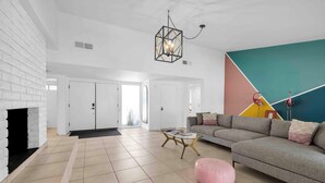 A spacious living room with a white brick fireplace, a modern chandelier, a colorful wall mural, a grey sofa, and a pink ottoman. - Avery (Palm Springs)