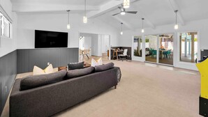 A modern living room with a large grey sofa, pendant lights, a ceiling fan, and a flat-screen TV mounted on the wall. - Avery (Palm Springs)