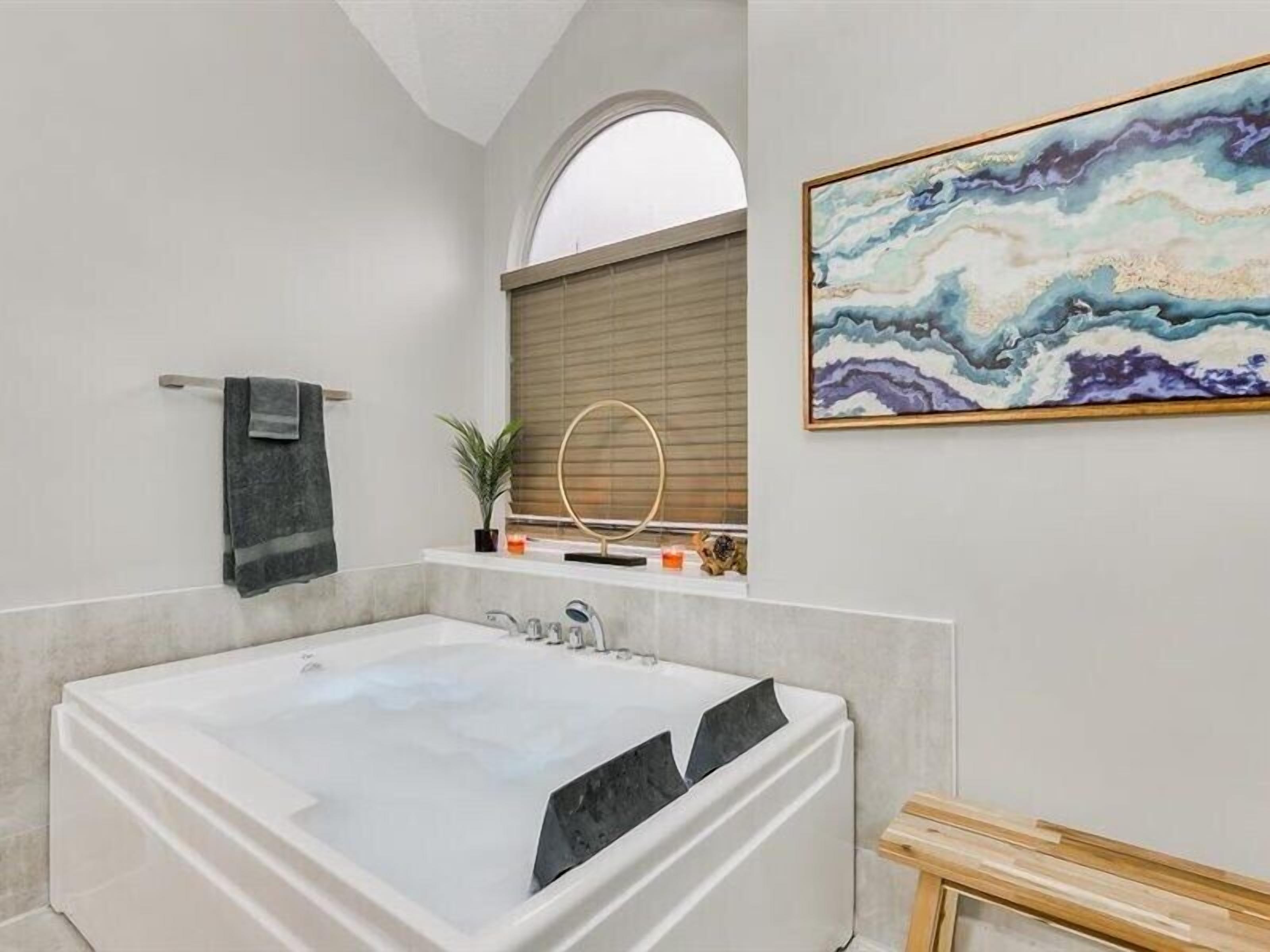 Een moderne badkamer met een groot bad, een handdoekhouder, een raam met zonwering en een schilderij aan de muur.
