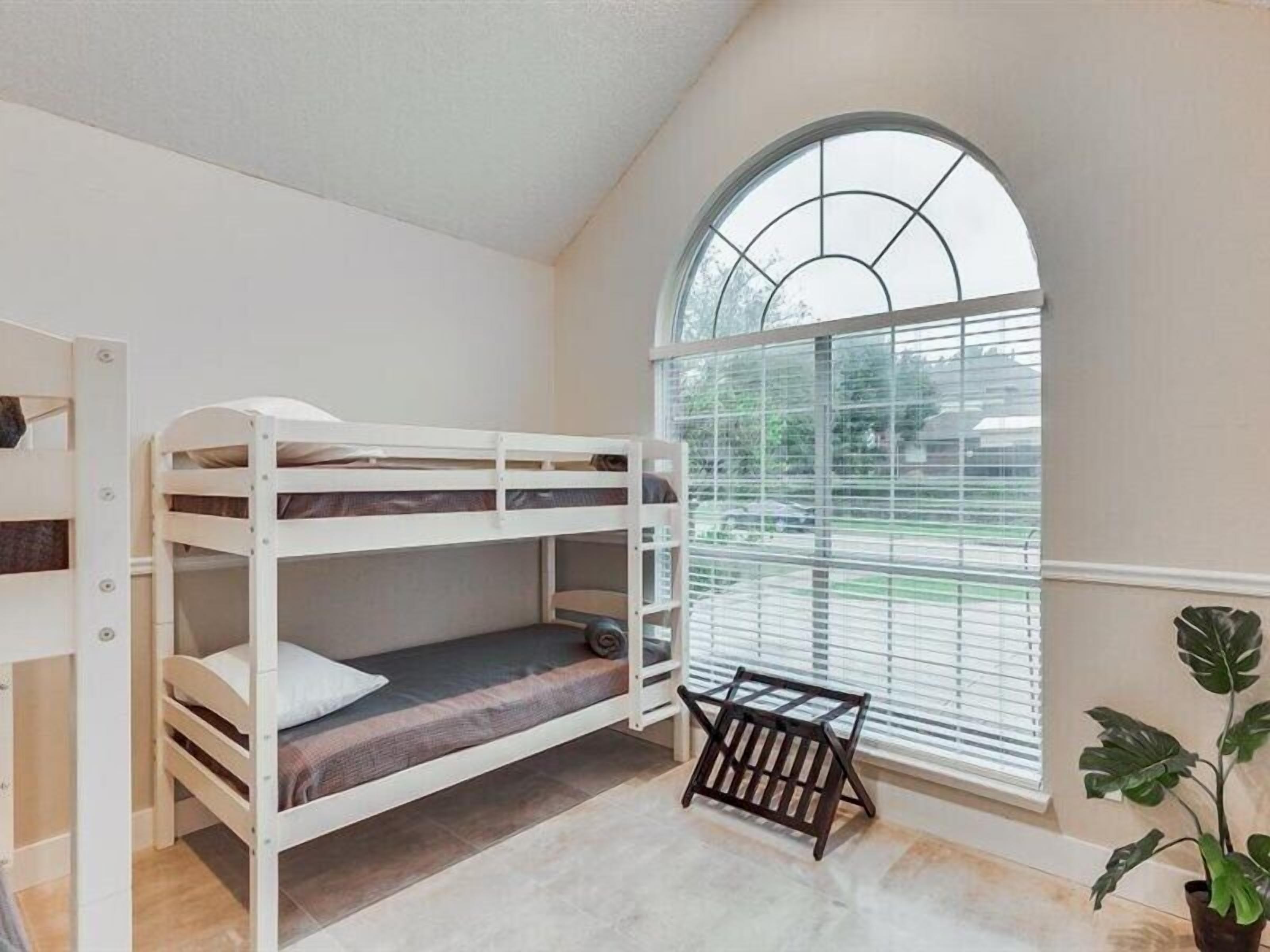 Een slaapkamer met stapelbed, een groot rondboogvenster, een tafeltje en een potplant.
