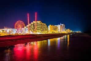 Une vue nocturne d’une ville, avec des bâtiments illuminés, une grande roue et une rivière qui reflète les lumières.