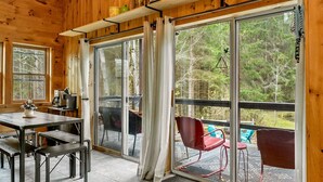 L’intérieur d’un chalet en bois comprend un coin repas, une table avec des chaises, un canapé et une porte-fenêtre coulissante qui donne sur une terrasse extérieure également équipée de chaises.