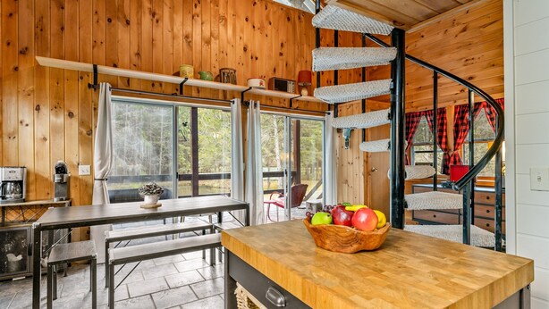 L’intérieur d’un chalet en bois comprend un coin repas, un comptoir de cuisine avec un panier de fruits et un escalier en colimaçon.
