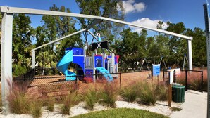 Un'area giochi con uno scivolo blu e strutture per arrampicarsi, circondata da una recinzione e alberi.