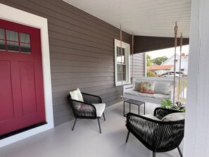 Un porche abrité avec une porte rouge, deux fauteuils en osier noirs, une petite table et un banc blanc avec des coussins.