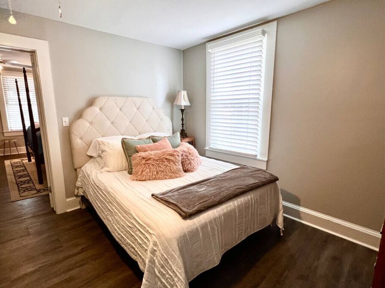 A bedroom with a tufted headboard, a bed with multiple pillows, a bedside lamp, and a window with blinds.