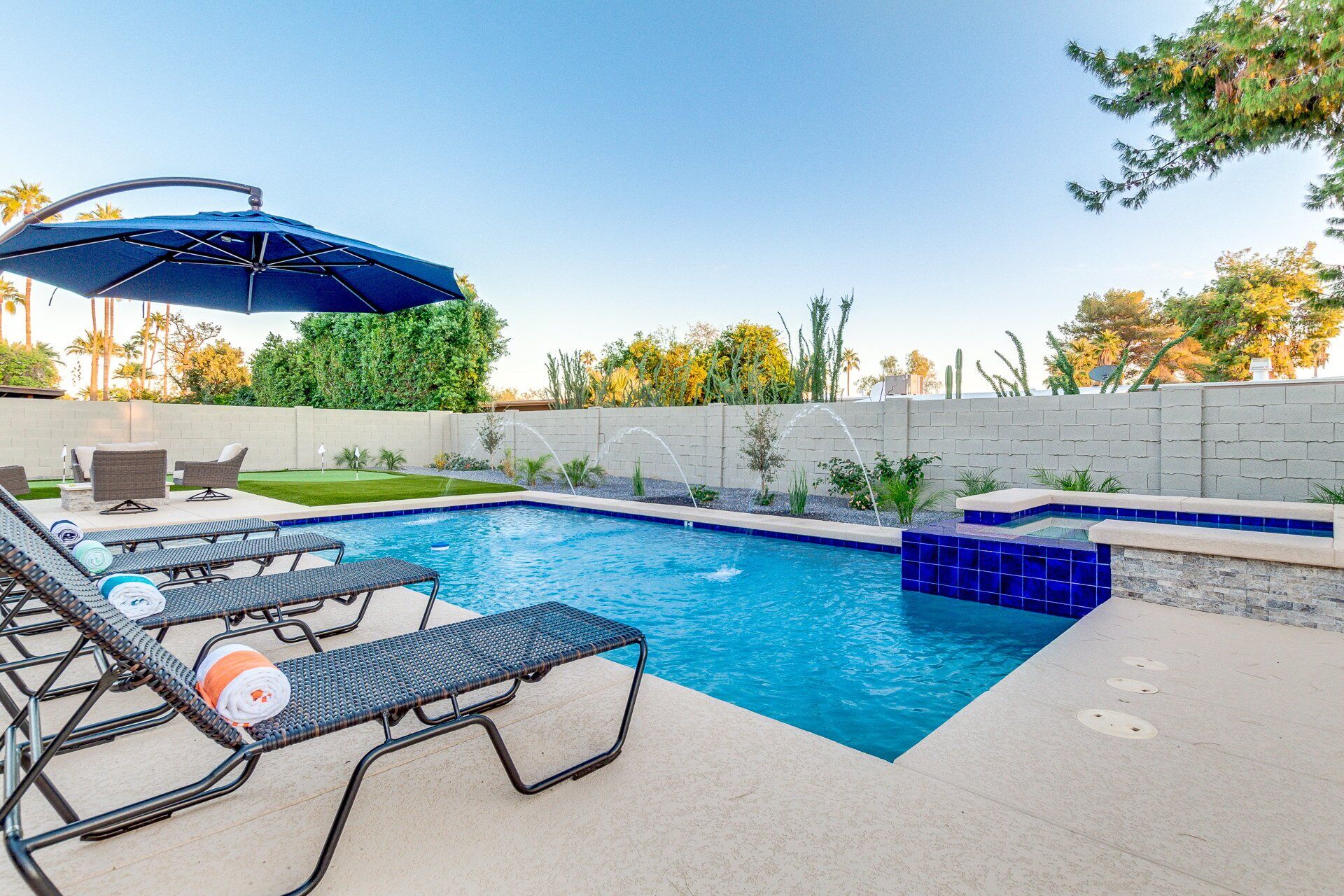 A swimming pool with lounge chairs and a blue umbrella, surrounded by a landscaped garden and a brick wall.