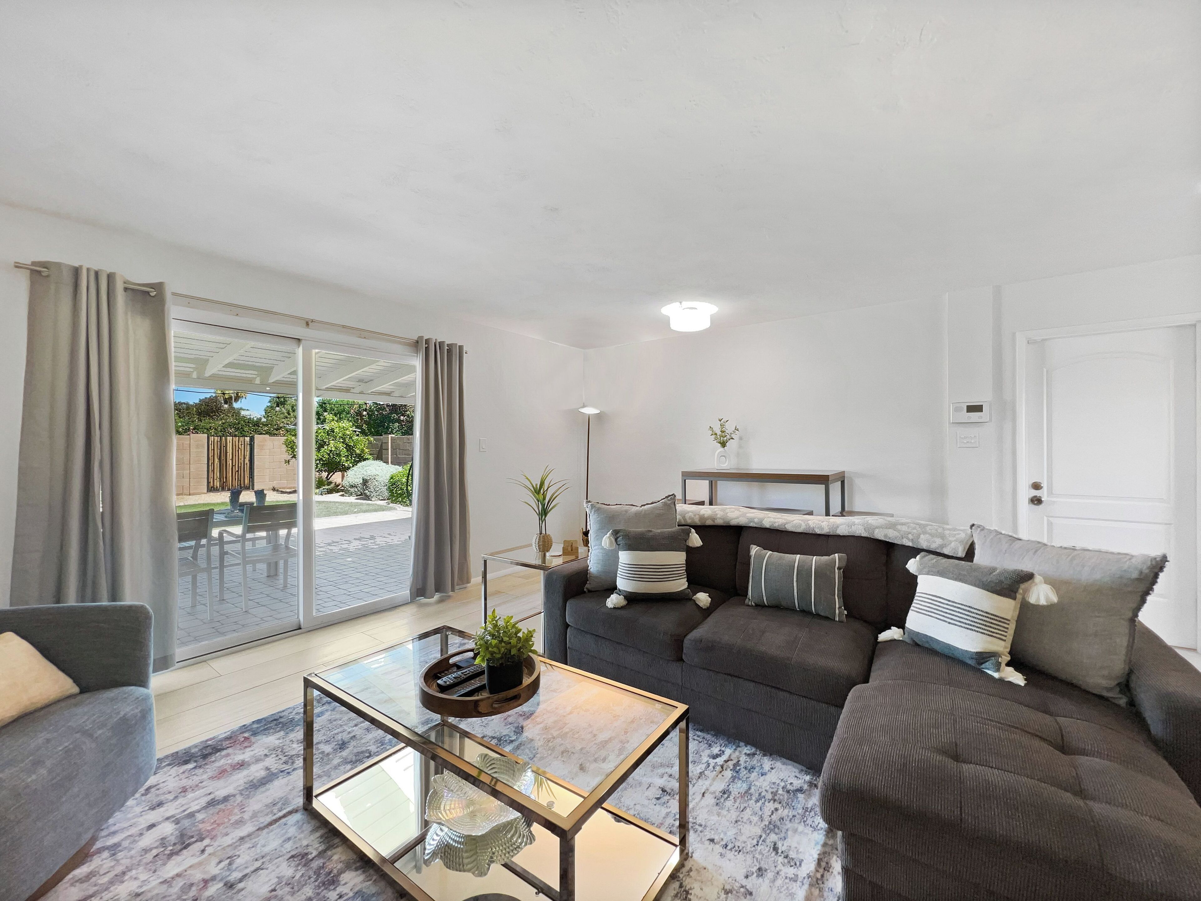 A modern living room with a glass coffee table, grey sofa, and a sliding glass door leading to a patio.
