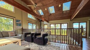 Ein geräumiges Zimmer mit hohen Decken, Holzbalken und Dachfenstern. Es gibt einen Balkon mit Geländer, eine Couch und zwei Sessel.