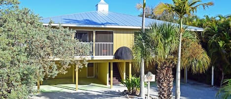 A yellow house with a blue roof, surrounded by palm trees and shrubs.