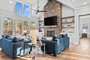 A spacious living room with a stone fireplace, large windows, and a flat-screen TV. - Lakeside Haven With Gourmet Kitchen (Hartwell)
