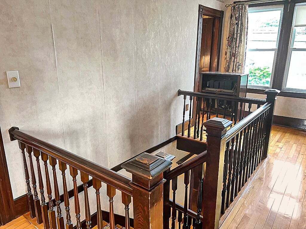 A wooden staircase with a handrail, a light switch on the wall, and a window with curtains.