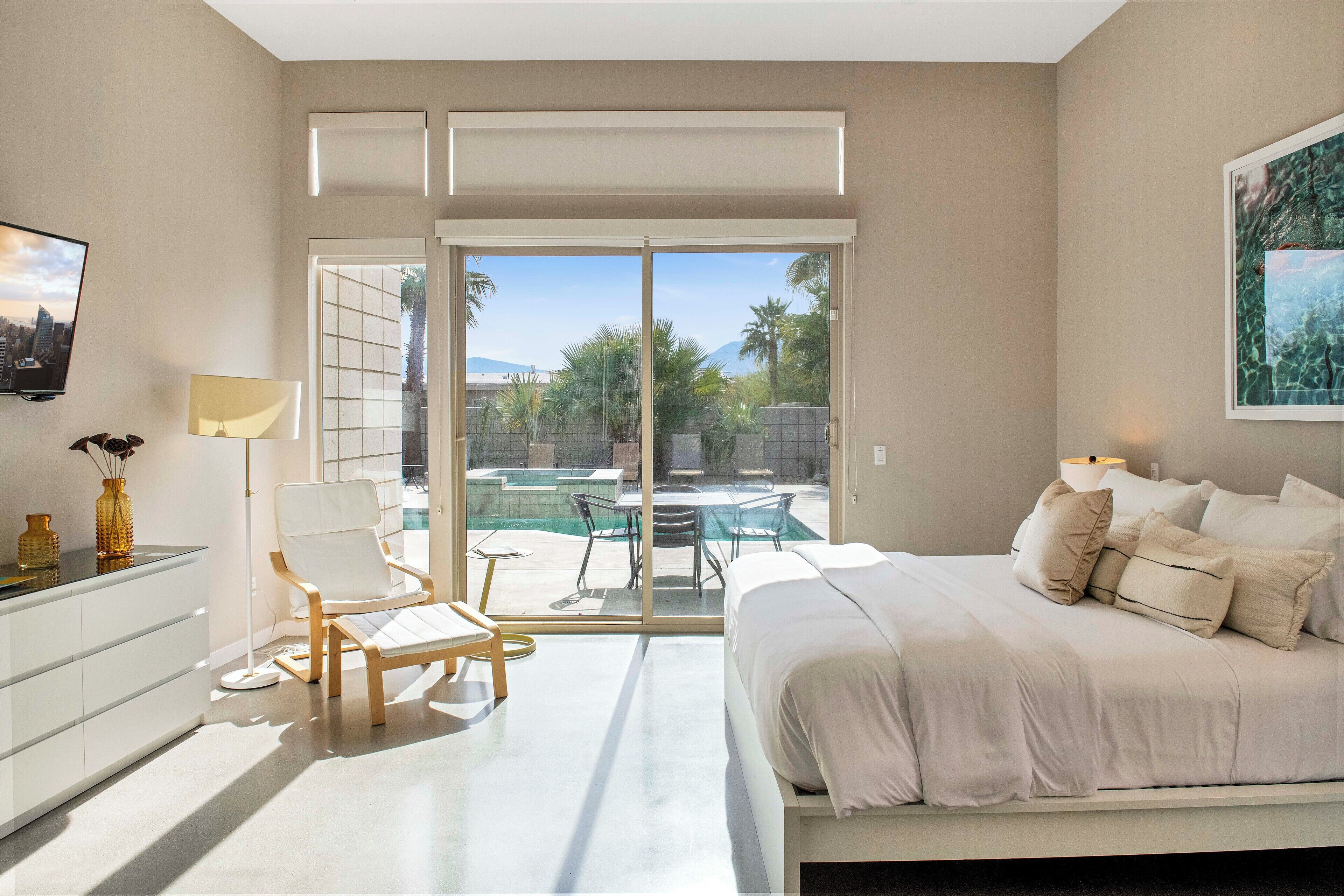 A bedroom with a bed, a chair, a desk, a TV, and a view of an outdoor pool and palm trees.