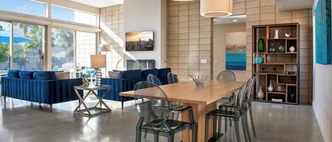A spacious living area with a large wooden dining table, clear glass chairs, and a blue sofa.