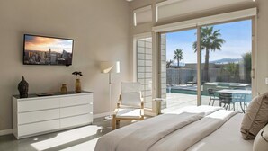 A modern bedroom with a bed, a white dresser, a chair, a TV, and a balcony with a pool and palm trees. - Lucent (Palm Springs)