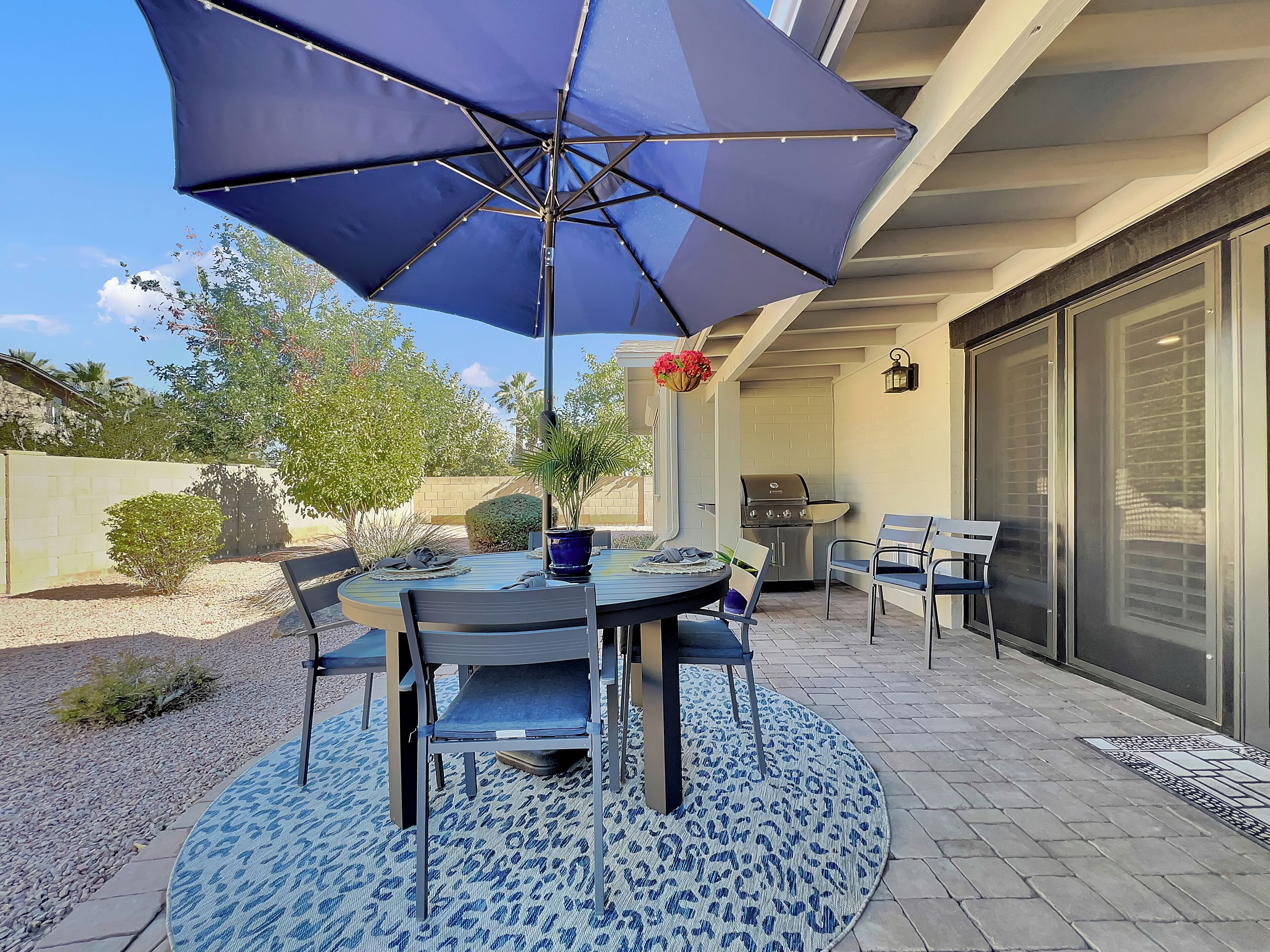 Une terrasse avec une table ronde, des chaises, un barbecue et un grand parasol.