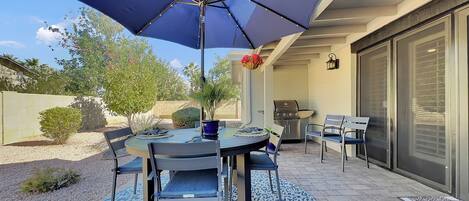 Une terrasse avec une table ronde, des chaises, un barbecue et un grand parasol.