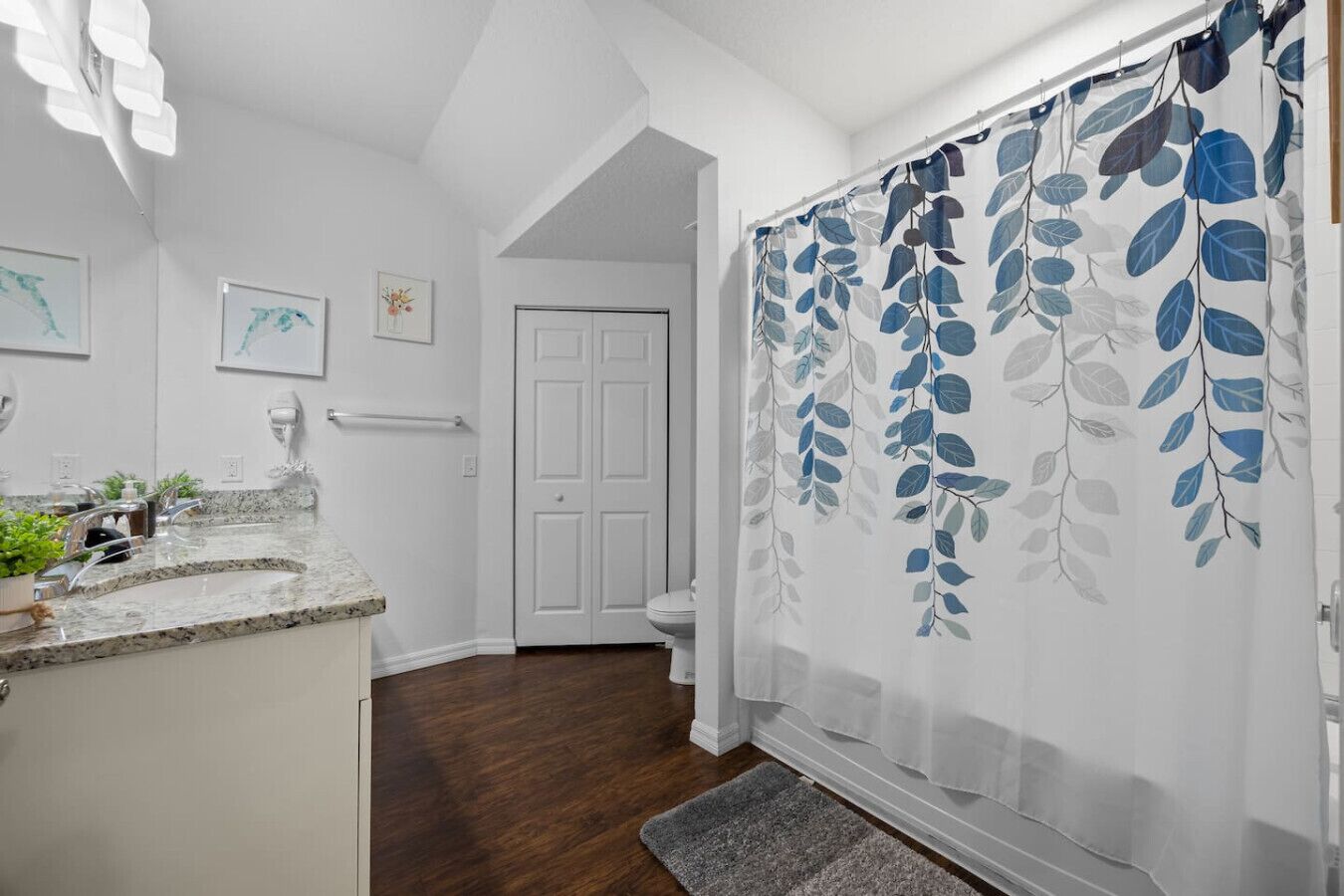 Un baño con tocador blanco, cortina de ducha con diseño de hojas y puerta que da a otra habitación.