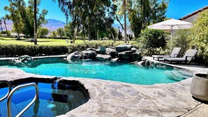 A swimming pool with a stone border, surrounded by rocks and greenery, with a view of mountains in the background.
