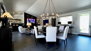 A modern dining area with a round table, white chairs, and a chandelier. There is a television mounted on the wall and a bar area in the background.