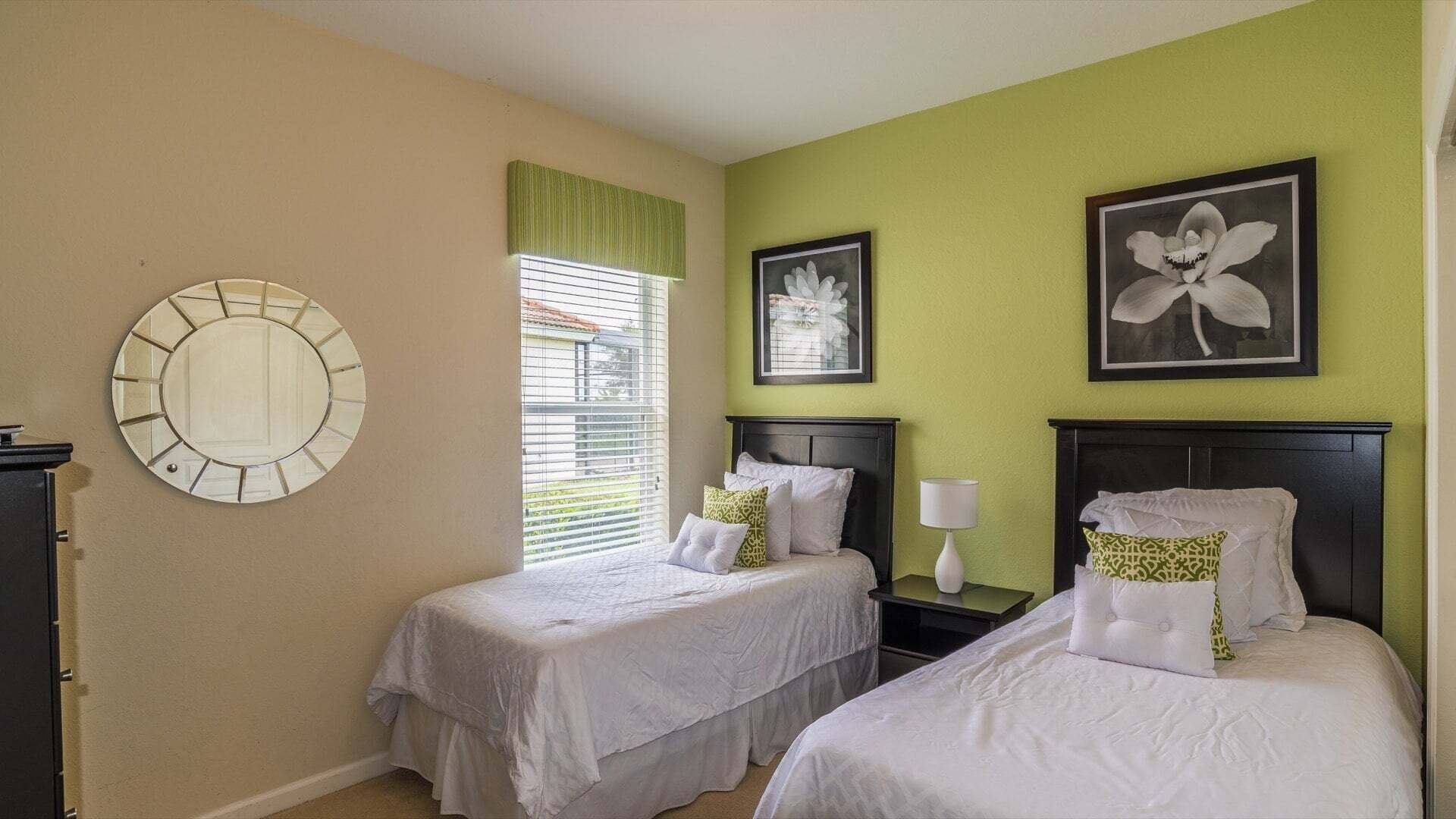 A hotel room with two beds, a round mirror, a window with blinds, and two framed pictures on the wall.
