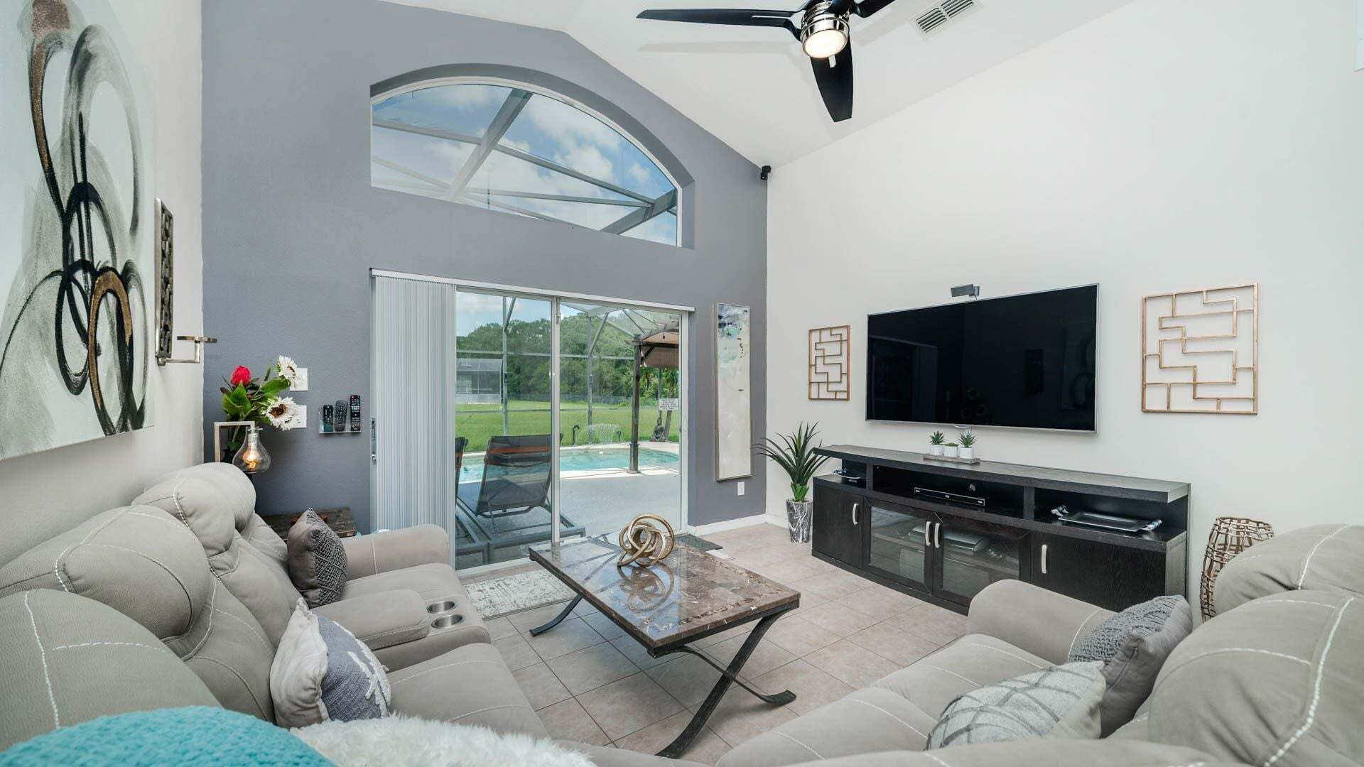 A modern living room with a large TV, comfortable seating, a glass door leading to a patio, and a ceiling fan.