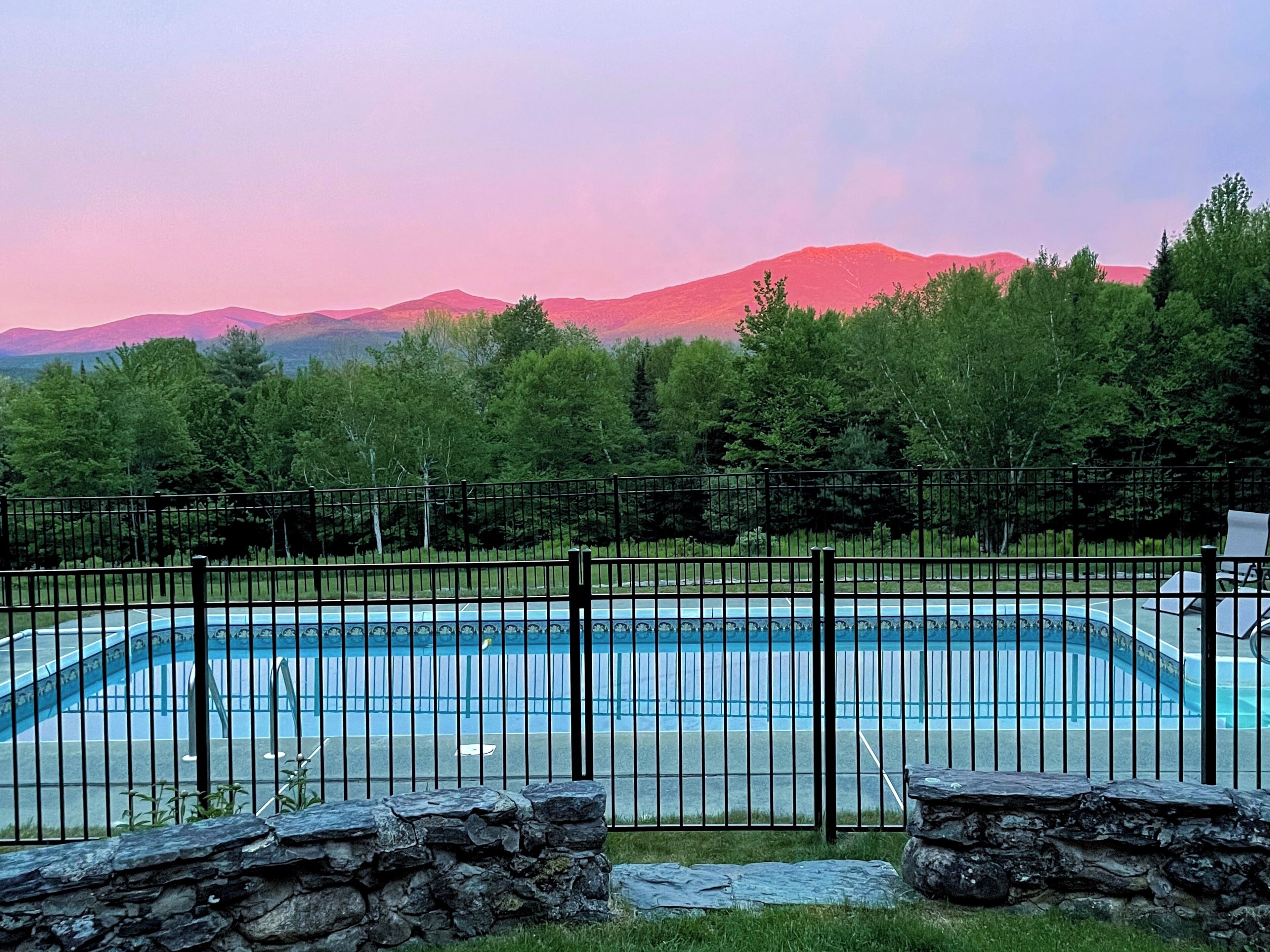 A swimming pool with a stone border, surrounded by a metal fence, and set against a backdrop of trees and mountains.