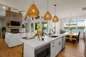 A modern kitchen with a large island, white cabinetry, and a stone fireplace. There is a dining area with a wooden table and chairs, and a view of the outdoors through large windows. - Hogan Crest (Mooresville)