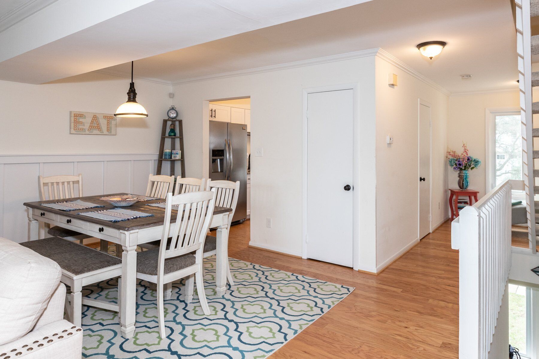 A dining area with a wooden table, white chairs, a patterned rug, and a refrigerator in the background.