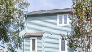 A two-story residential building with a garage, a small front yard, and a tree with pink flowers. - Latitude Adjustment (Virginia Beach)