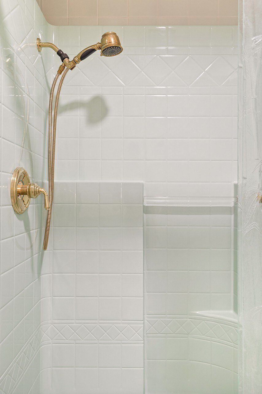 Une douche dotée d’un pommeau et d’un flexible dorés, de murs recouverts de carreaux blancs et d’un design d’angle.