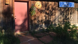 Un bâtiment rustique avec une porte en bois, une jardinière suspendue, un décor en forme de soleil et un panneau indicateur pointant vers « TRIMBLE HOT SPRINGS ».