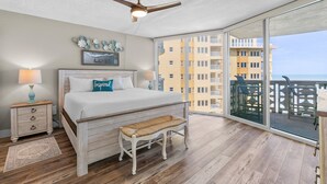 Een slaapkamer met een groot bed, een bankje, een plafondventilator en een balkon met uitzicht op de stad.