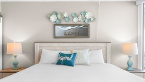 Een net opgemaakt bed met witte lakens, een sierkussen met de tekst 'Inspired' en een kunstwerk erboven.
