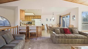 A spacious living area with a sofa, a coffee table, and a dining table with chairs. A kitchen is visible in the background with wooden cabinets and a refrigerator. - Fairway Village 8 (Bend)