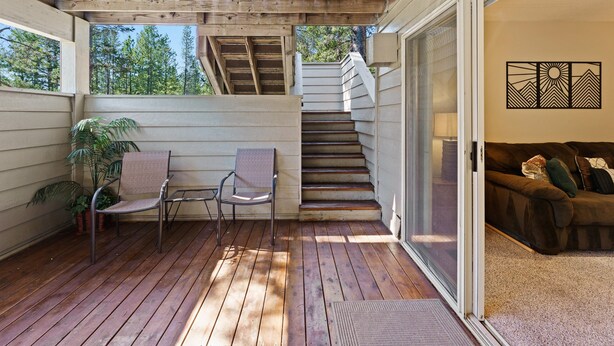 Une terrasse en bois avec une chaise et un pot de fleurs, un escalier menant à un deuxième niveau, et une porte-fenêtre coulissante donnant sur un salon avec un canapé.