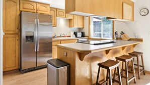 Une cuisine moderne avec des meubles en bois, un réfrigérateur en acier inoxydable, un îlot central intégré avec des tabourets et une fenêtre laissant entrer la lumière naturelle.