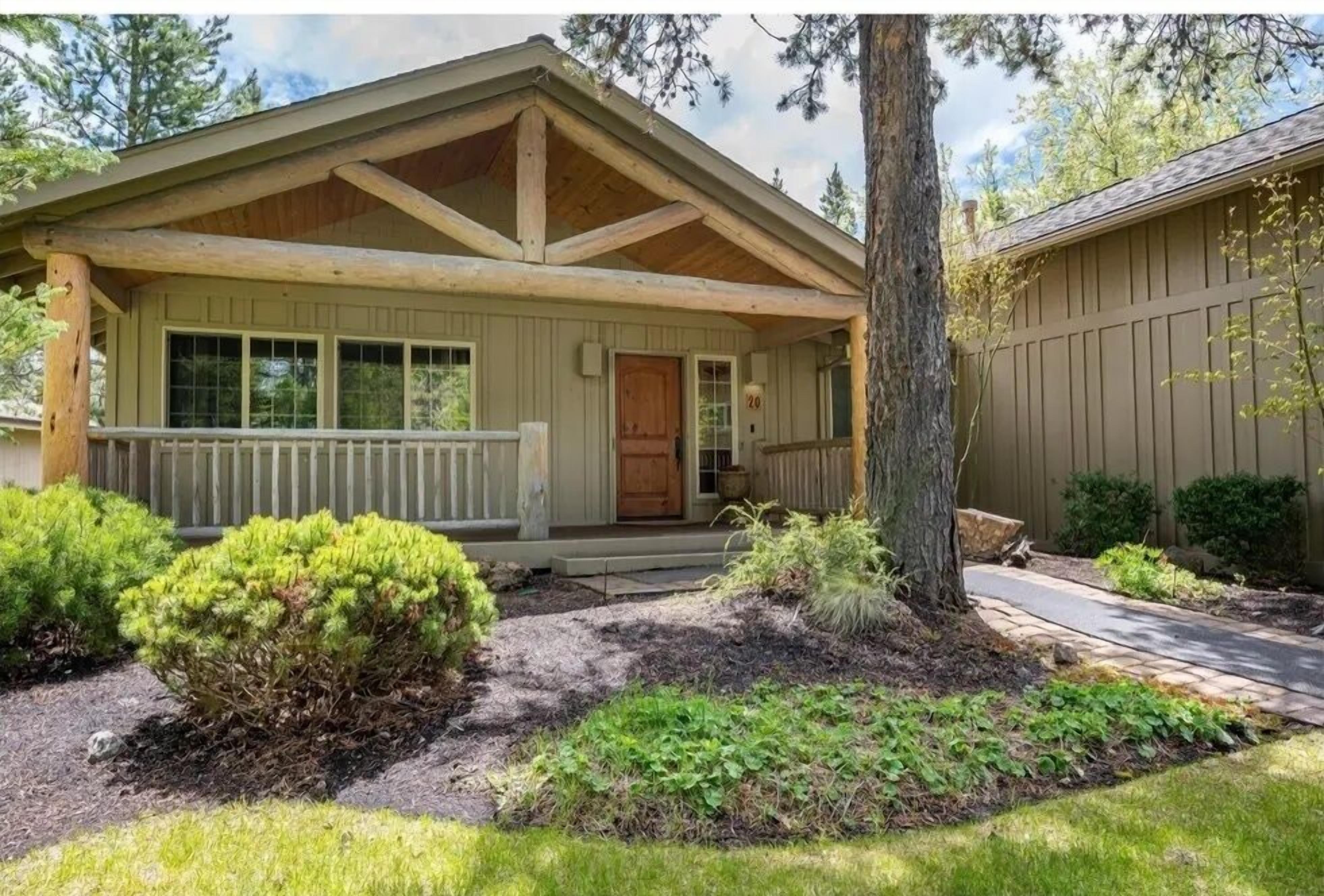 A single-story house with a front porch, a wooden deck, and a well-maintained garden with various shrubs and a tree.