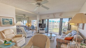 A spacious living room with a large TV, comfortable seating, and a balcony with a view of the city. - Errol By The Sea 406 (New Smyrna Beach)