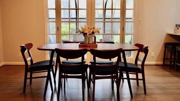 Un coin repas avec une table et des chaises en bois, un vase de fleurs et une vue dégagée sur l’extérieur grâce à de grandes fenêtres.