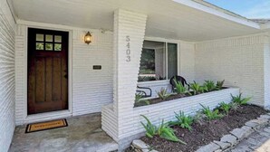 Une maison en briques blanches avec une porte d’entrée marron foncé, le numéro « 5403 » inscrit sur le mur, et un petit jardin avec des plantes.