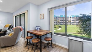 Une chambre moderne équipée d’une table à manger, de tabourets de bar, d’un fauteuil de massage et d’une grande fenêtre donnant sur un bâtiment et un espace verdoyant.