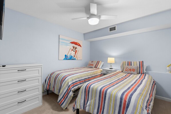 A bedroom with two beds, a dresser, a ceiling fan, and a painting on the wall. - Errol By The Sea 207 (New Smyrna Beach)